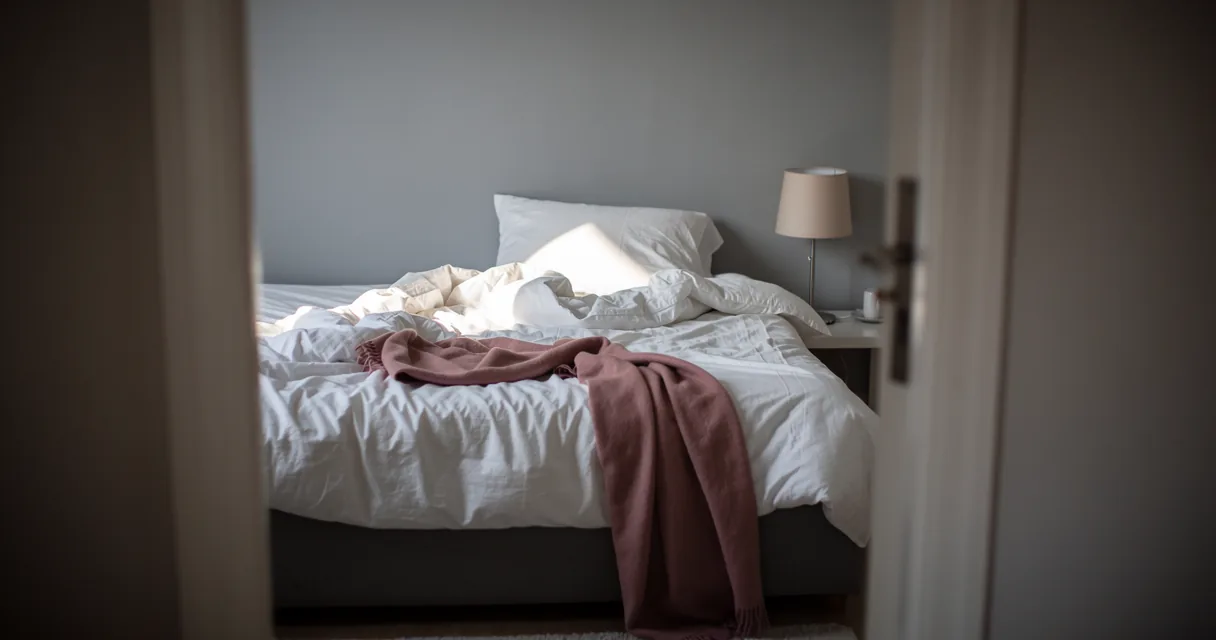 Morning light on a rumpled white bed with a rose blanket and a bedside lamp